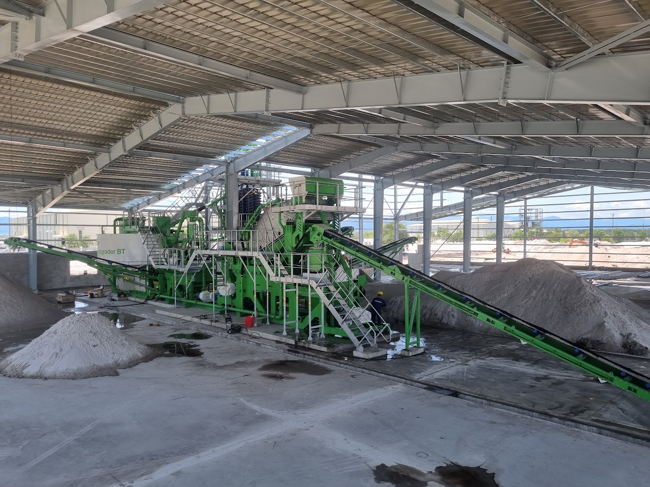 Large lime-green industrial hydrograder and conveyor system processing bulk material inside a warehouse