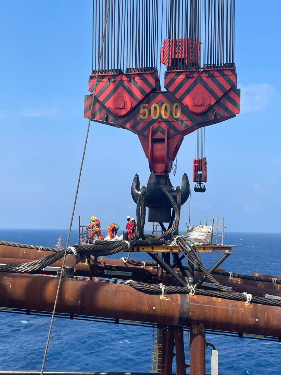 Crew rigging large steel pipes to a red 5000-tonne crane block over open water