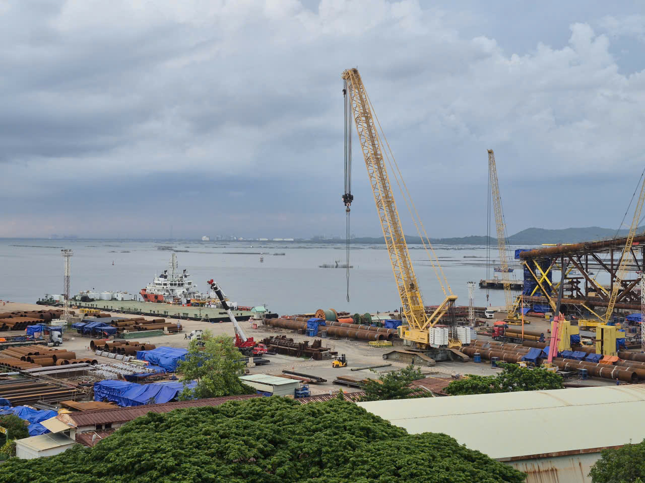 Coastal shipyard with yellow cranes, stacked pipe, and offshore jacket structures under cloudy skies