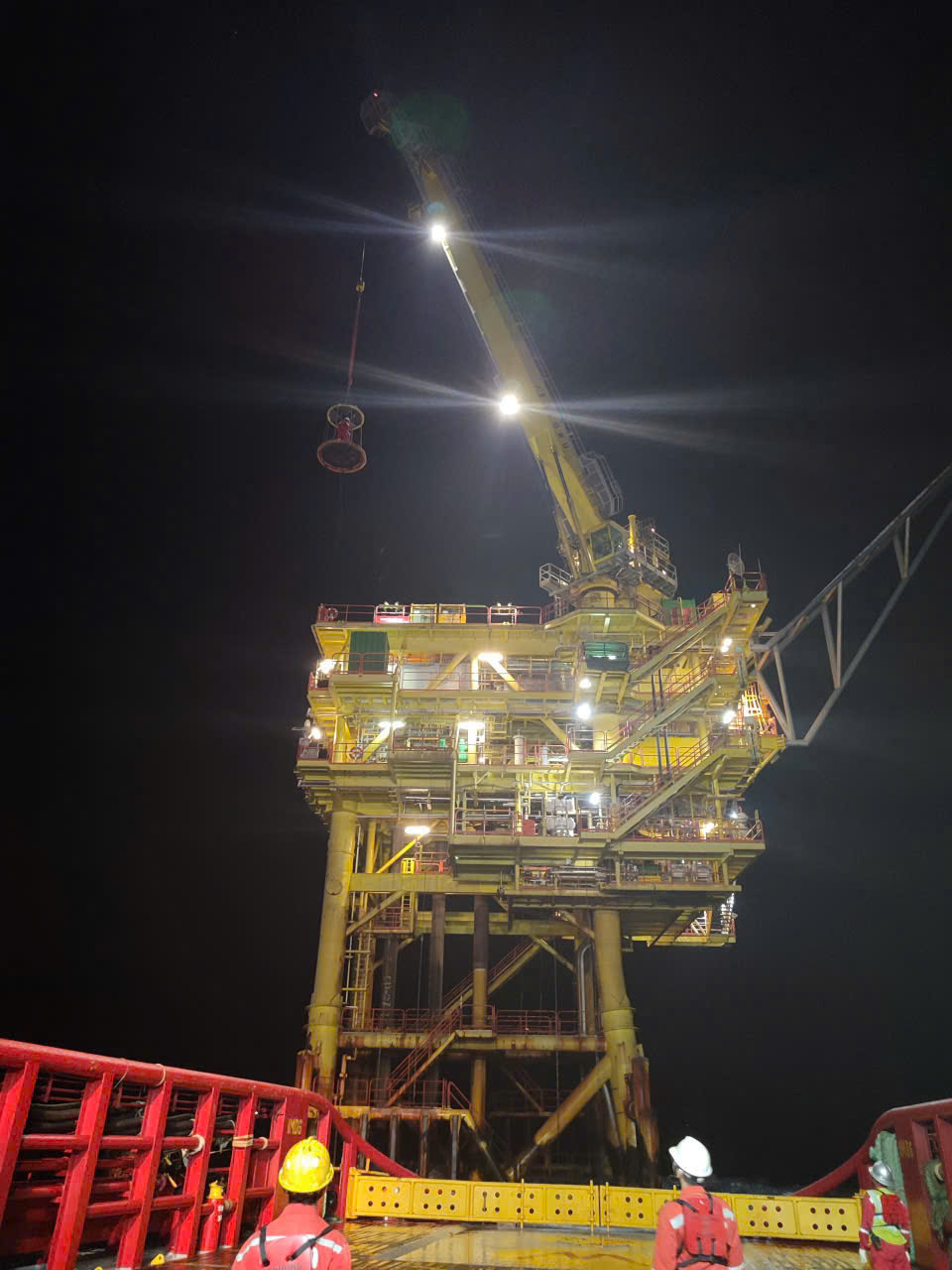 Nighttime offshore platform lit by floodlights with crew on a support vessel and personnel transfer basket